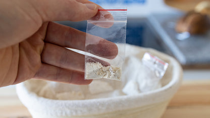 A hand holding a sealed pack of dehydrated Explorer Starter flakes above a proofing basket